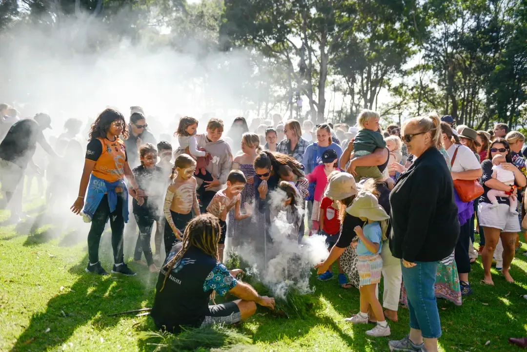A diverse crowd, including children, gathers outdoors for a smoke ceremony, with sunlight filtering through the trees.