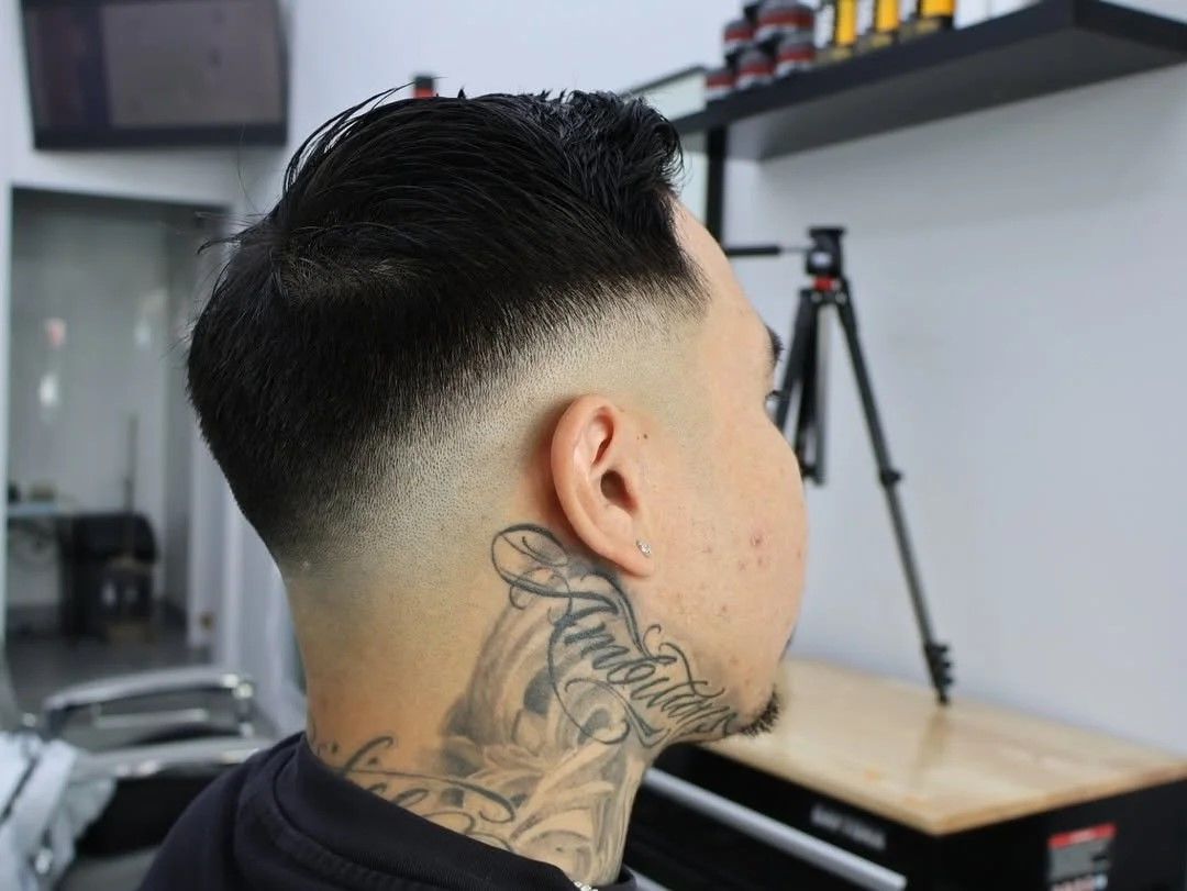 Person with a fresh fade haircut, tattooed neck featuring script text, seated in a modern barbershop with a tripod and shelves in the background.