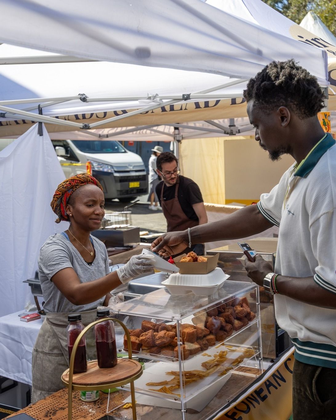A woman serves food from a stall at an outdoor market to a man. A staff member works in the background. There are fried snacks and beverages on display.