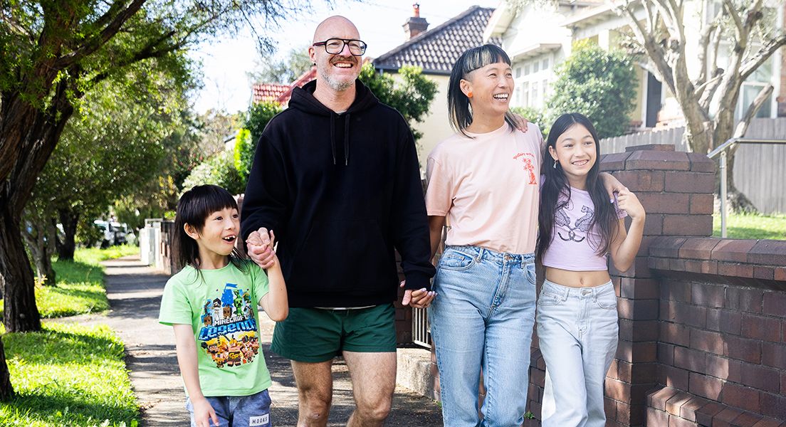 A family of four, smiling and holding hands, walks along a suburban sidewalk lined with trees and brick fences on a sunny day.