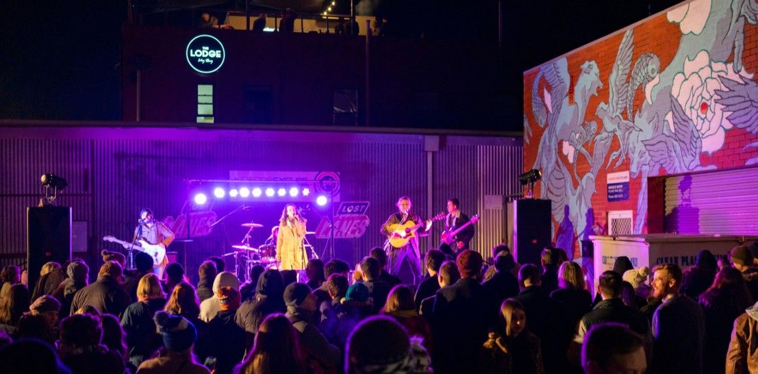 Outdoor night concert with a band performing on stage, colorful lights, a mural on a nearby wall, and a crowd gathered in front.