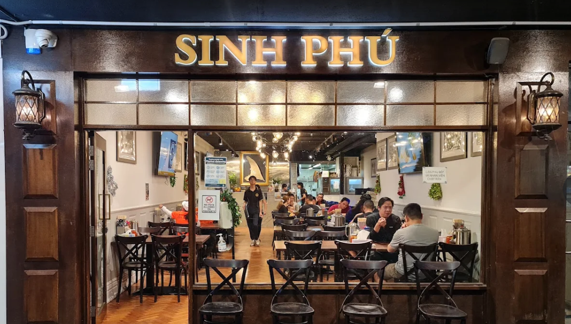 A cosy restaurant named "Sinh Phú" with wooden decor, filled with people dining at tables. Warm lighting and wall art create a welcoming atmosphere.