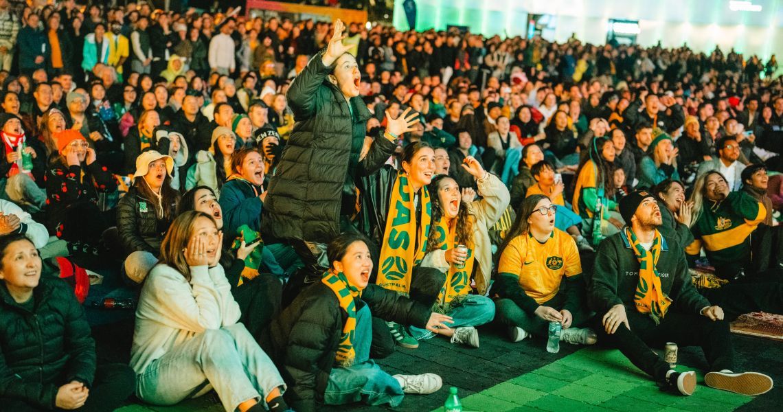 A large crowd, many wearing yellow, watches an event on a big screen, displaying varied expressions of excitement and anticipation.