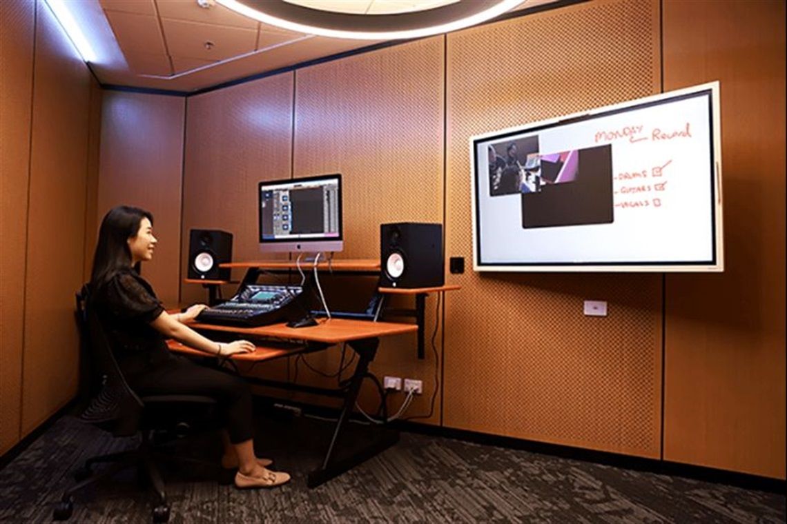 A woman sits at a desk with a computer and speakers in a modern recording studio, facing a large wall-mounted screen displaying notes.