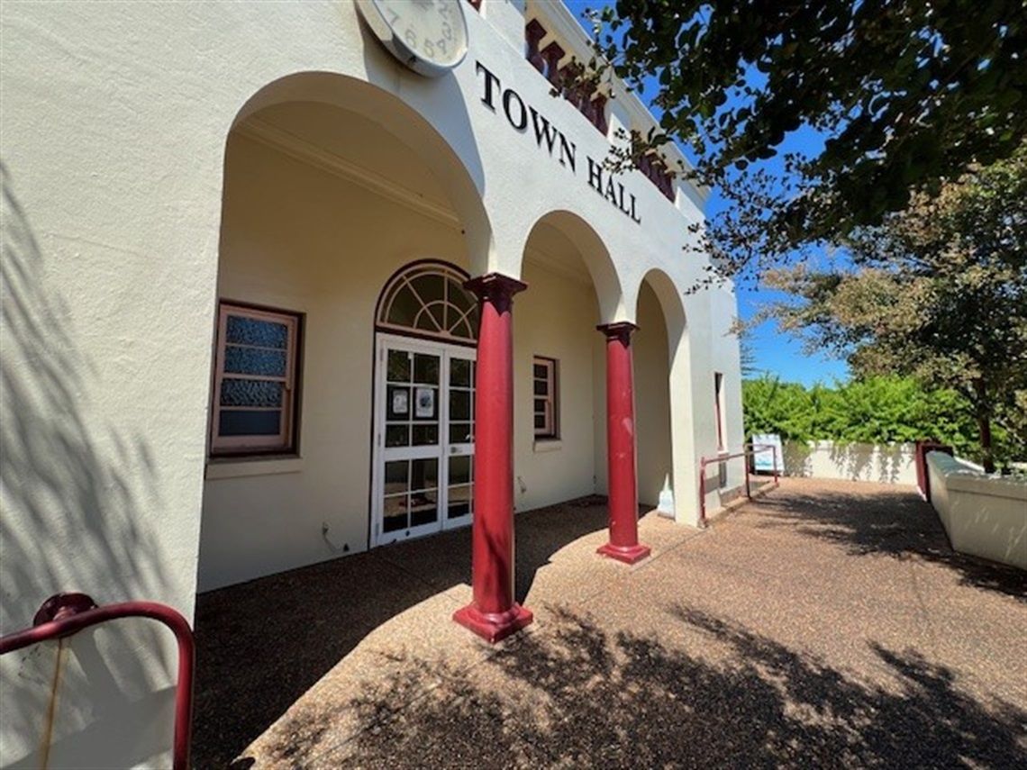Entrance of a town hall with arched doorways, red columns, and a pathway lined with trees under a clear blue sky.