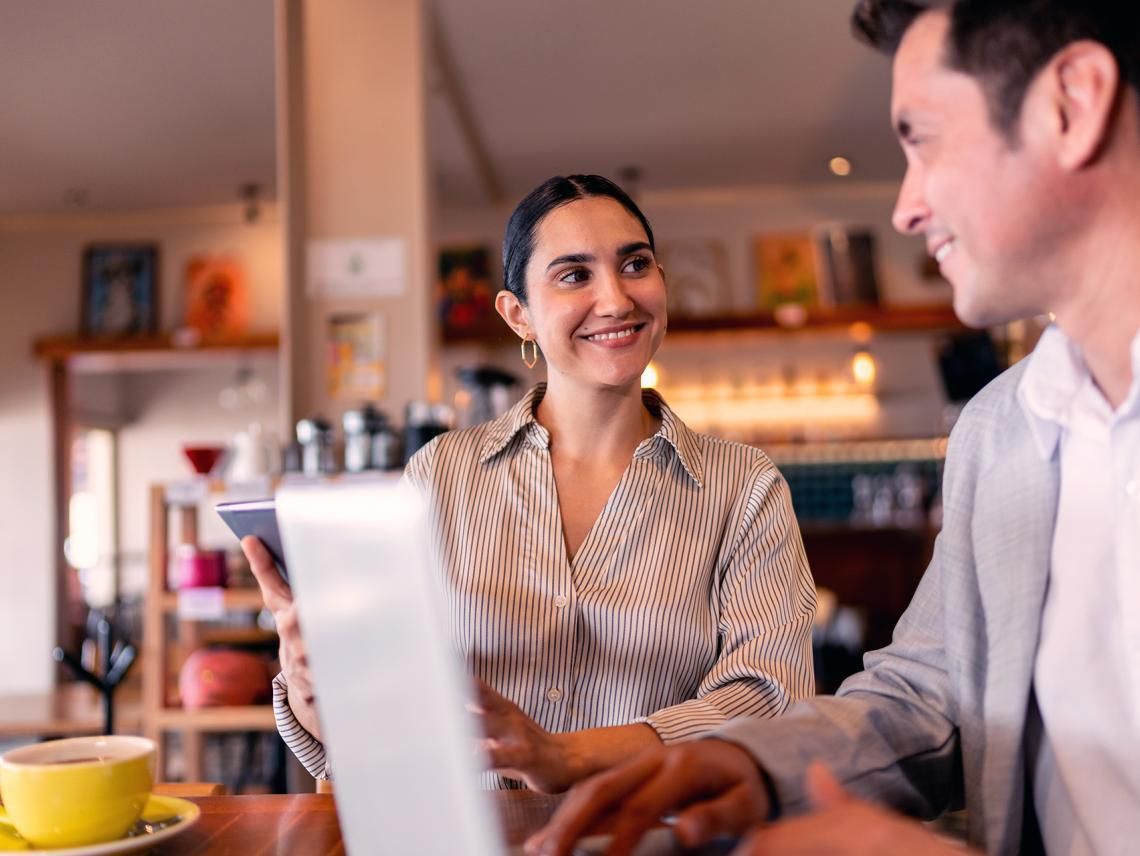 Two people sitting in a cafe, smiling at each other. One holds a tablet while the other appears to be using a laptop.