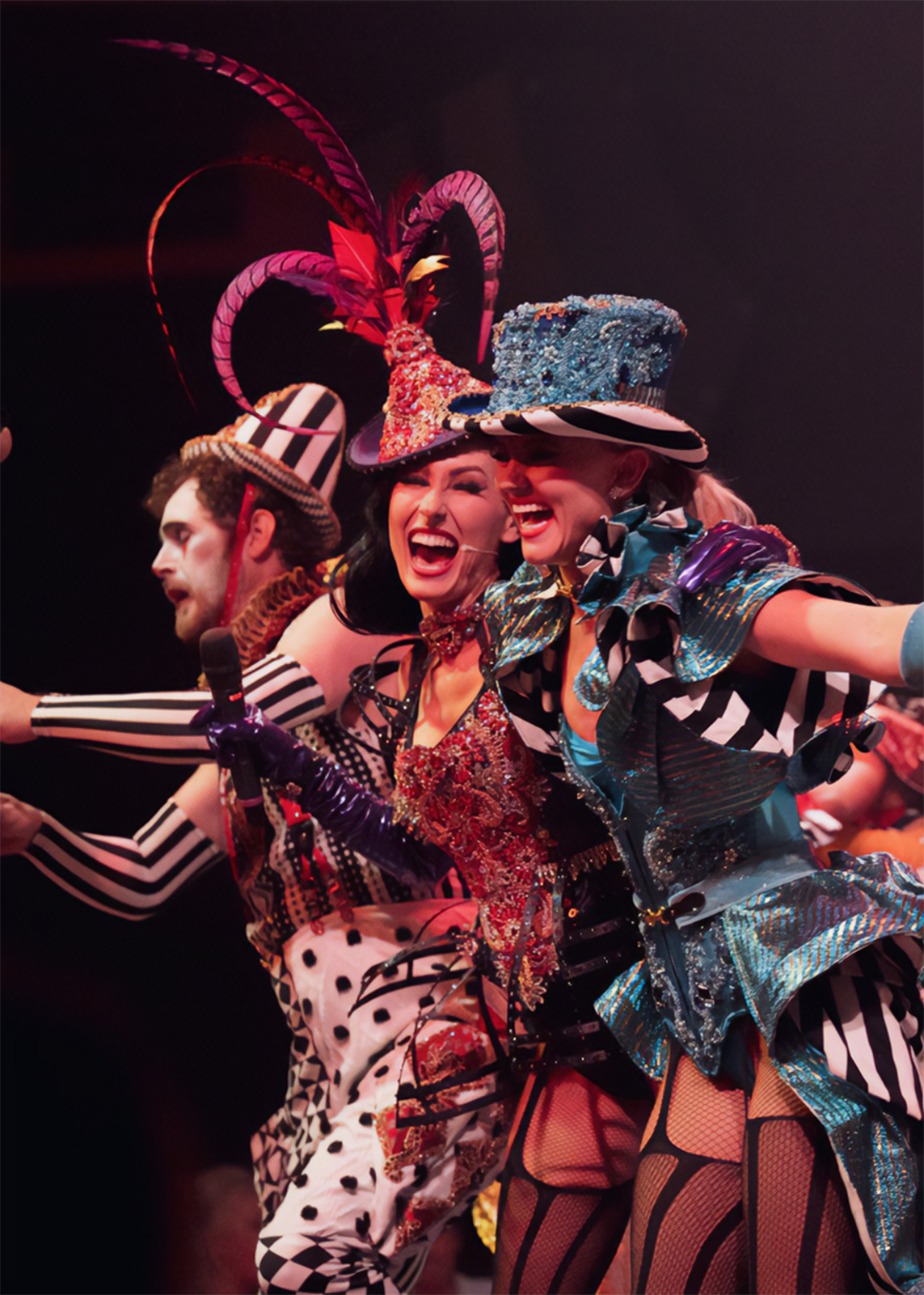 Three theatrical performers captured mid-finale on stage in an explosion of elaborate costuming and pure joy. Centre frame, a female performer in a heavily jewelled crimson and black harlequin-patterned corset throws her head back laughing, wearing an ornate rhinestone-encrusted miniature top hat adorned with sweeping magenta feathers and curved horns. To her right, a second female performer beams broadly in a structured teal and black geometric costume with layered ruffled panels and a jewelled powder-blue top hat. To the left, a male performer in monochrome black-and-white striped attire and a matching striped tall hat leans into the group mid-song. All three are pressed together in a moment of shared elation, arms wide and faces fully alive. The background is dark and warm, drawing all attention to the richly saturated costumes and expressions. A theatrical lighting design by Lightology Australia frames the performers in warm-toned stage light, sculpting each costume with depth and dimension against the dark stage.