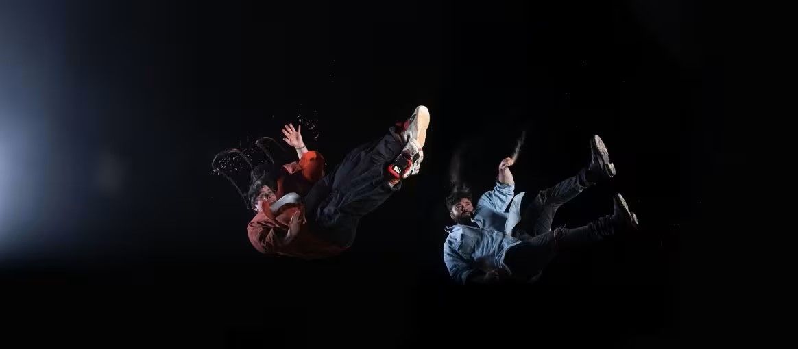 Two people in mid-air against a dark background, wearing casual clothes and sneakers, with light illuminating the left side.