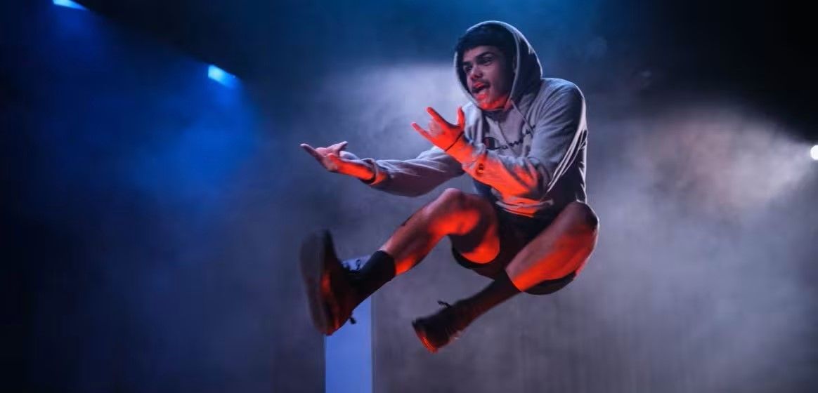 A dancer in a hoodie jumps energetically, with fog and blue lighting creating a dramatic stage effect.