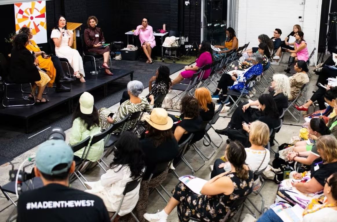 Audience attentively listens to a panel of speakers during an indoor event, with diverse participants seated in rows.