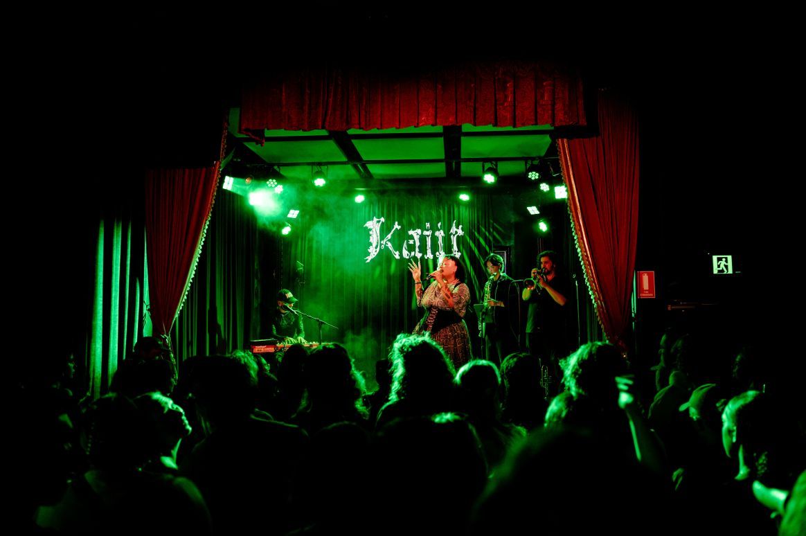 Band performs on a dimly lit stage with green lights and a red curtain backdrop, in front of a lively audience.