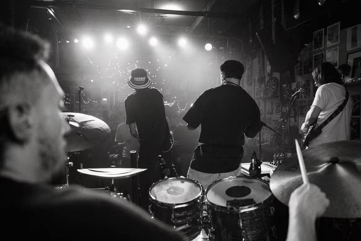 Black and white image of a band performing on stage, seen from behind the drummer. Audience and bright stage lights in the background.