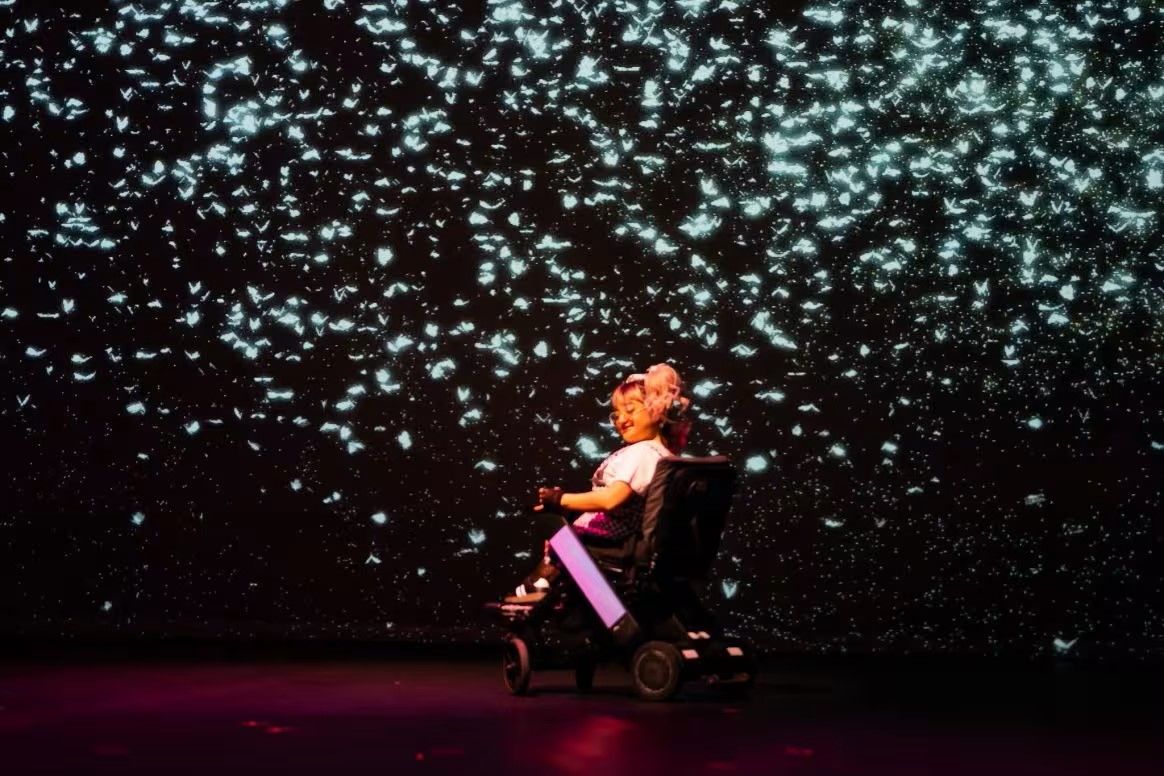 A person in a wheelchair is positioned on stage, surrounded by a dark background filled with glowing, scattered light patterns.