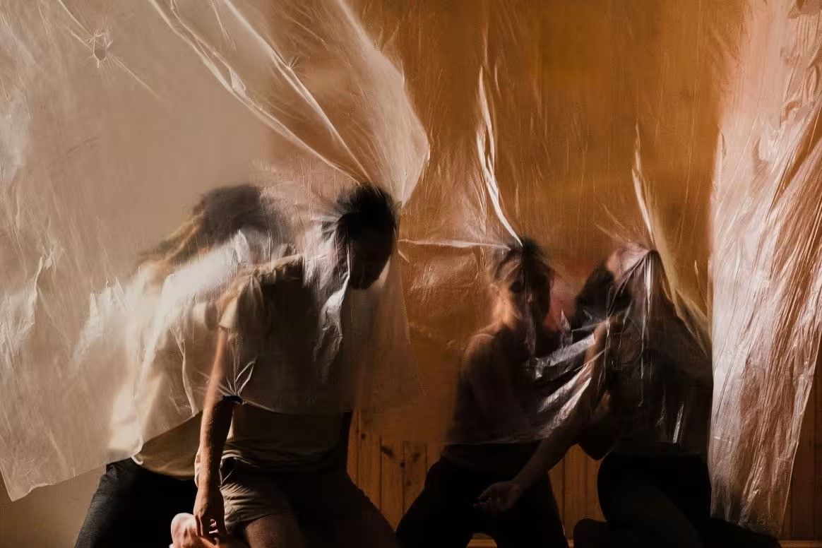 People sit beneath flowing translucent fabric, creating a blurred, dynamic effect against a warm, softly lit background.