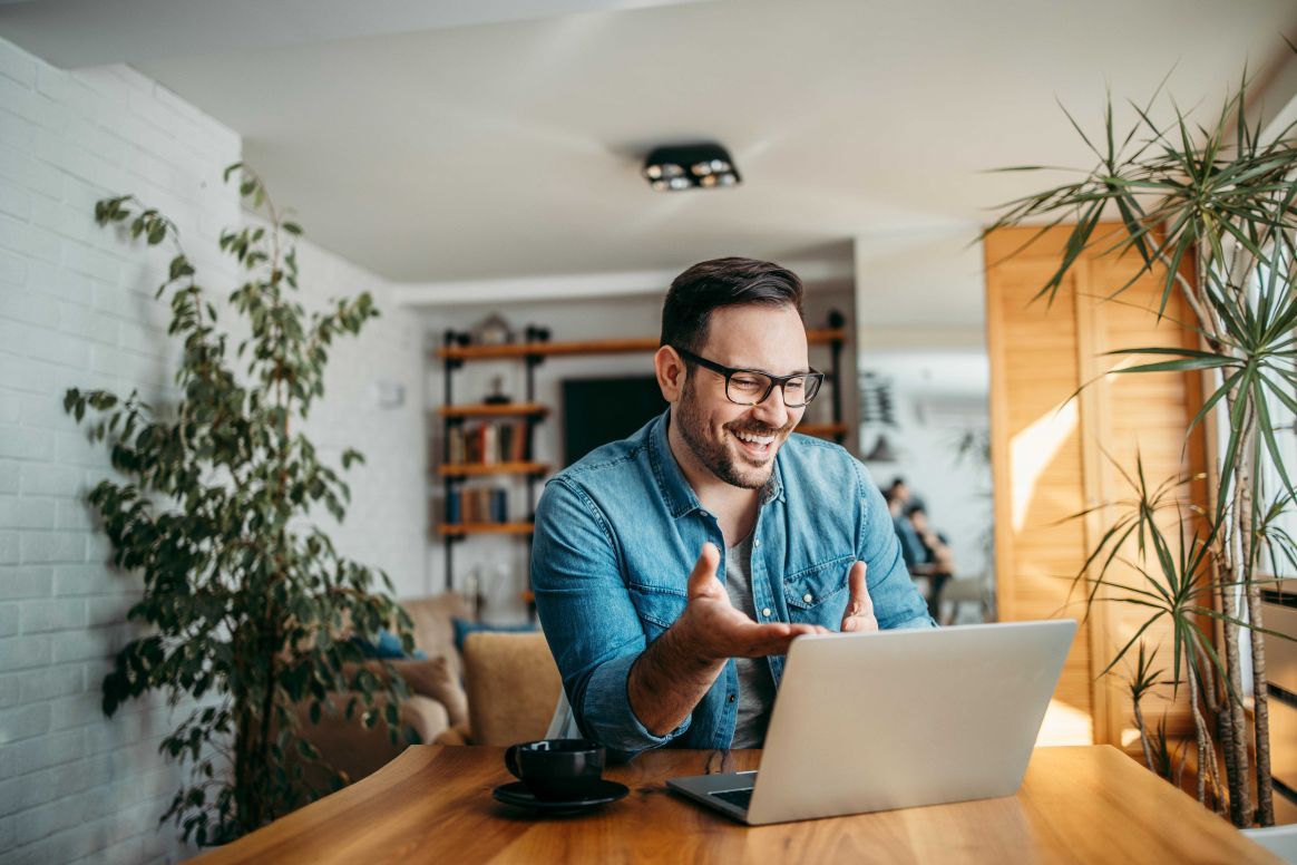 Man in glasses, smiling and gesturing, having a video call on a laptop at a wooden table, surrounded by plants in a cozy room.