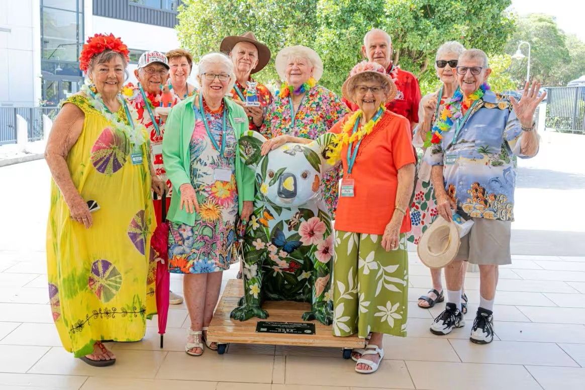 A cheerful group of older adults dressed in colourful Hawaiian attire poses around a decorative sculpture outdoors.