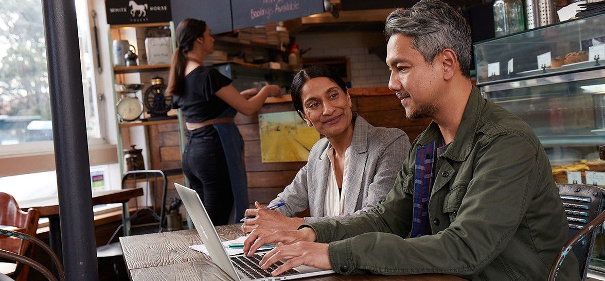 Two people sitting at a cafe table, with one using a laptop. A woman stands in the background preparing food.