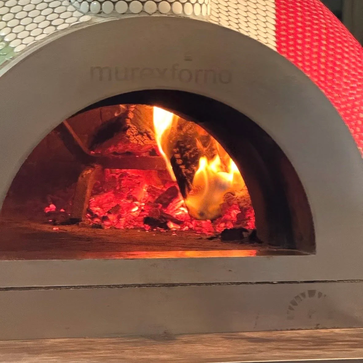 Wood-fired oven with a visible flame and embers, featuring a tiled archway exterior in white and red.