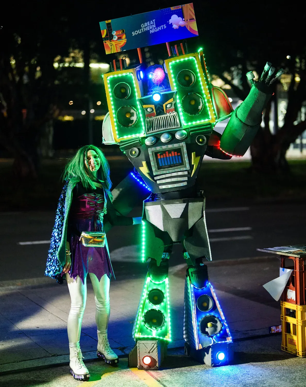 Person in a colorful costume poses next to a large, illuminated robot with speakers and a "Great Southern Nights" sign at night.