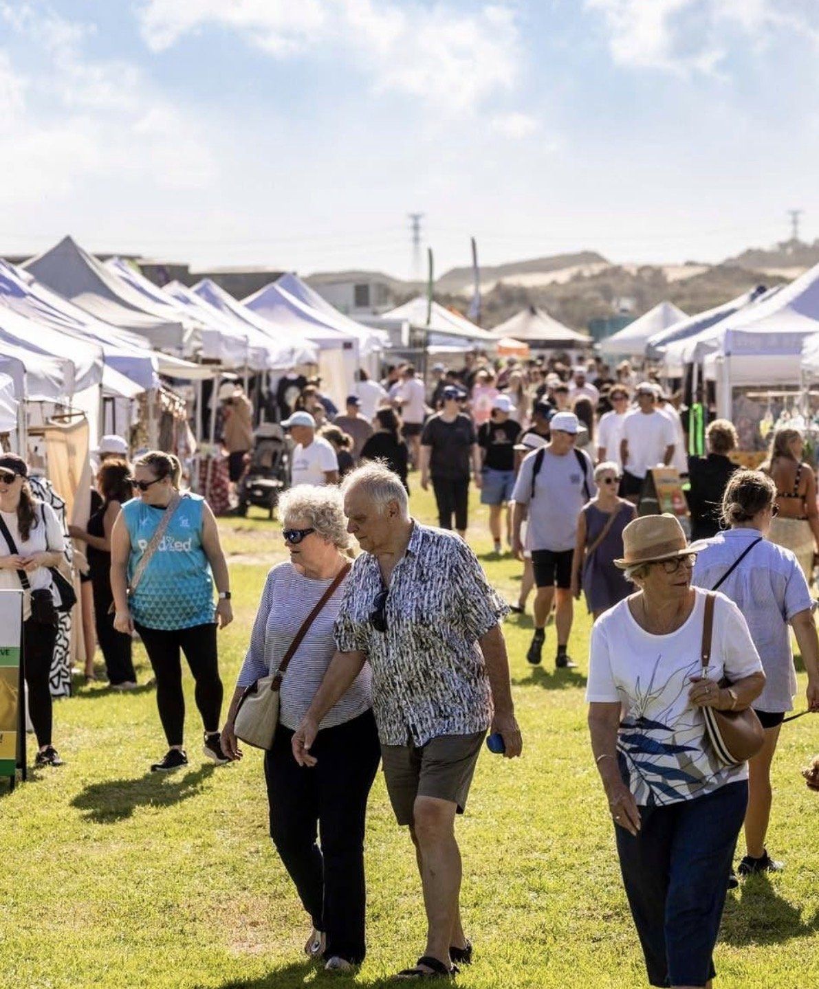 A bustling outdoor market with people walking between white-tented stalls on a sunny day, set against a backdrop of hills.