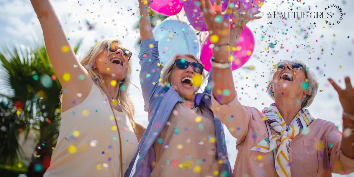 Three women joyfully throw confetti into the air, smiling and wearing sunglasses, with colorful balloons in the background on a sunny day.