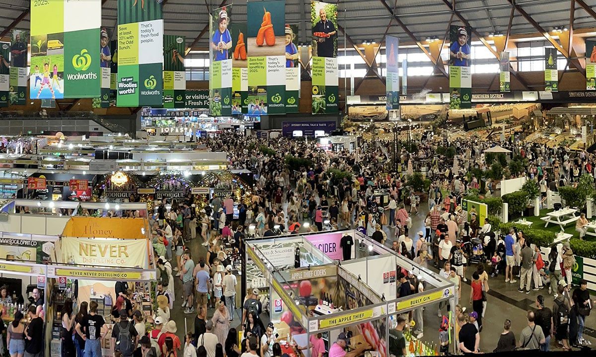 Crowded indoor exhibition space with numerous stalls and people walking around, featuring large overhead banners and a vibrant atmosphere.