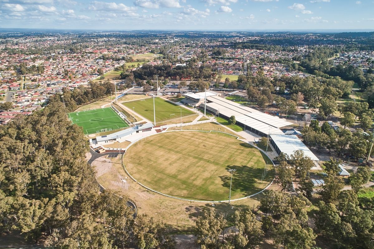 Aerial view of a sports complex with a large circular field, adjacent rectangular fields, surrounded by trees and residential areas.