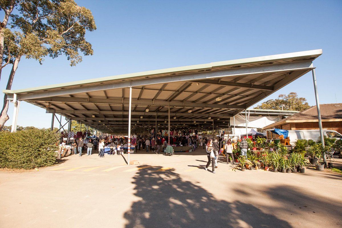 Outdoor market under a large metal canopy, with people browsing stalls and plants, surrounded by trees on a sunny day.