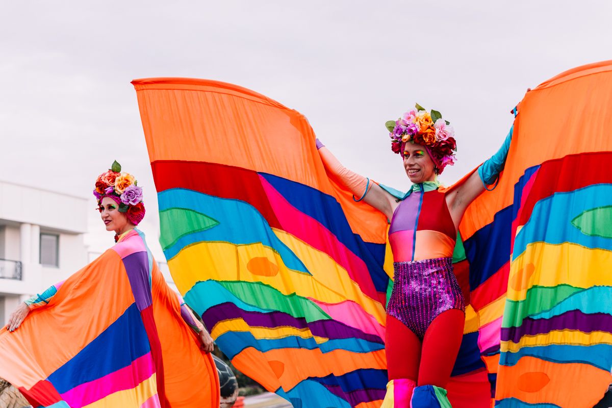 Two performers in vibrant, colorful costumes with large, wing-like fabric extensions and flower crowns, pose outdoors against a cloudy sky.