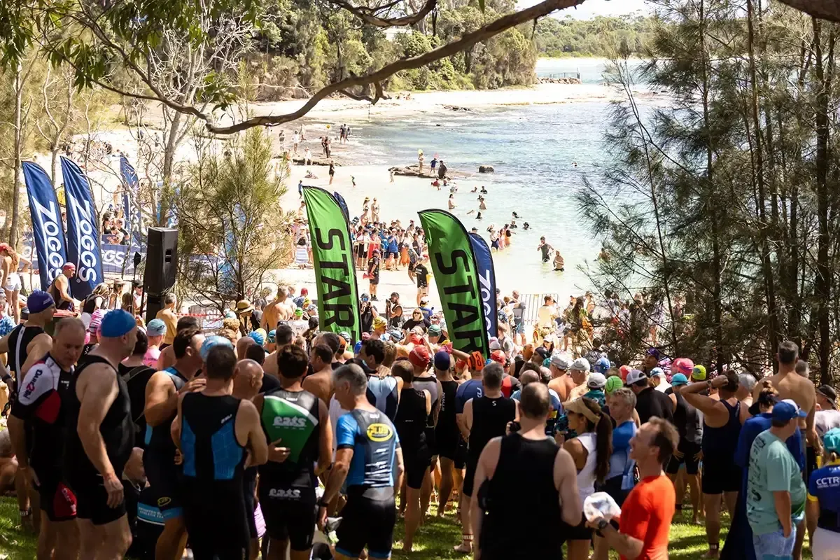 Crowd of swimmers gather at a beach start line for a race, surrounded by green "START" flags, trees, and onlookers in the background.