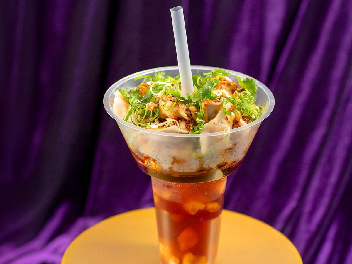 A drink cup with a straw, topped with colourful dumplings, garnished with greens and served on an orange surface against a purple background.