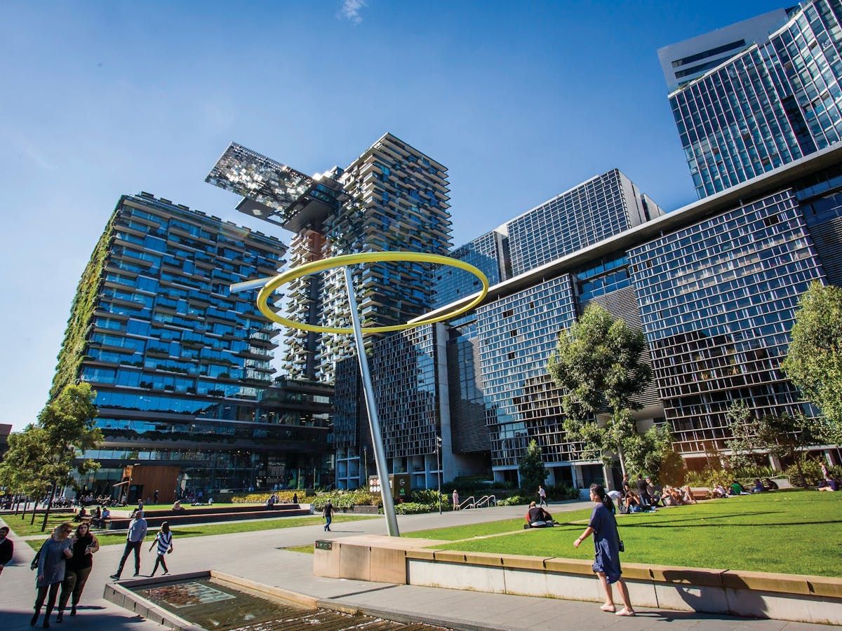 Modern urban park with a ring sculpture, lush greenery, and innovative glass architecture. People are relaxing under a clear blue sky.