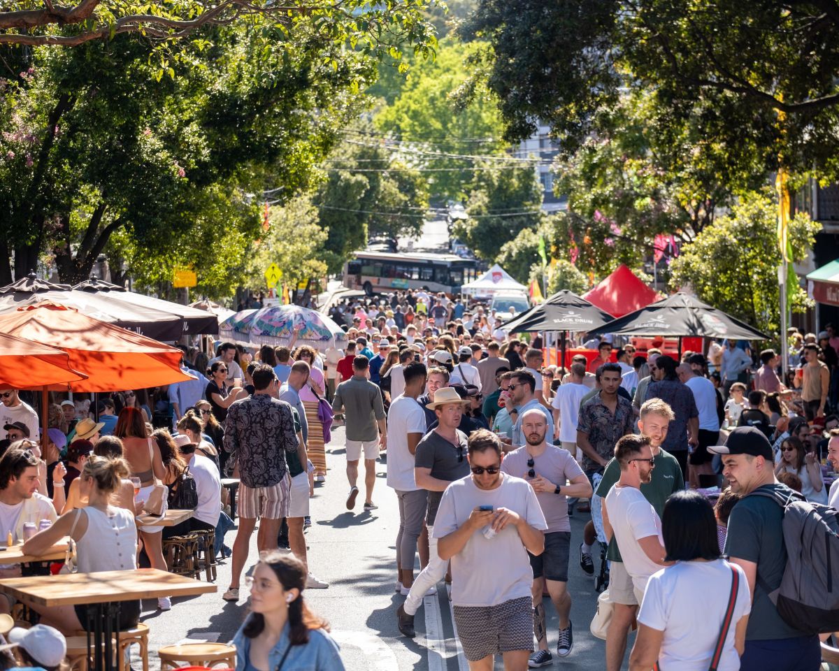 A lively street festival with a crowd of people enjoying the sunny day, surrounded by trees and colorful market stalls.