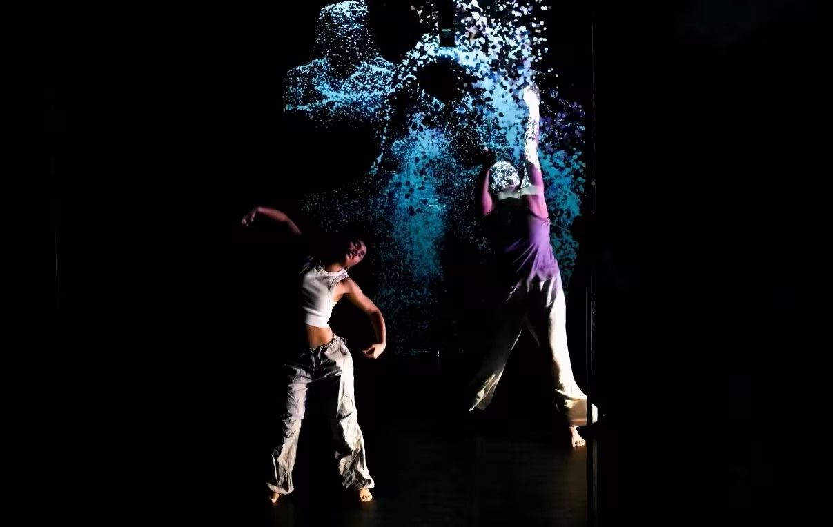 Two dancers perform dynamically on stage, one reaching upwards, with blue and white digital art projected in the background.