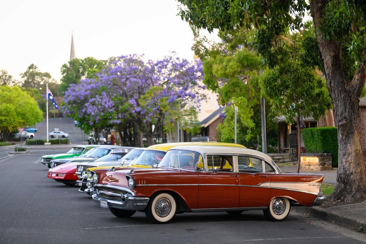 A row of classic cars, including a brown vintage car in the foreground, parked on a street lined with trees and purple flowering jacarandas.