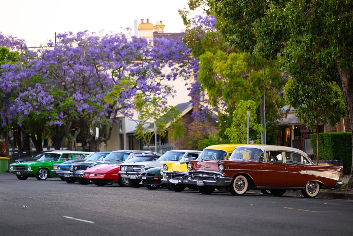 A row of colorful vintage cars parked on a street lined with jacaranda trees in bloom, with buildings in the background.
