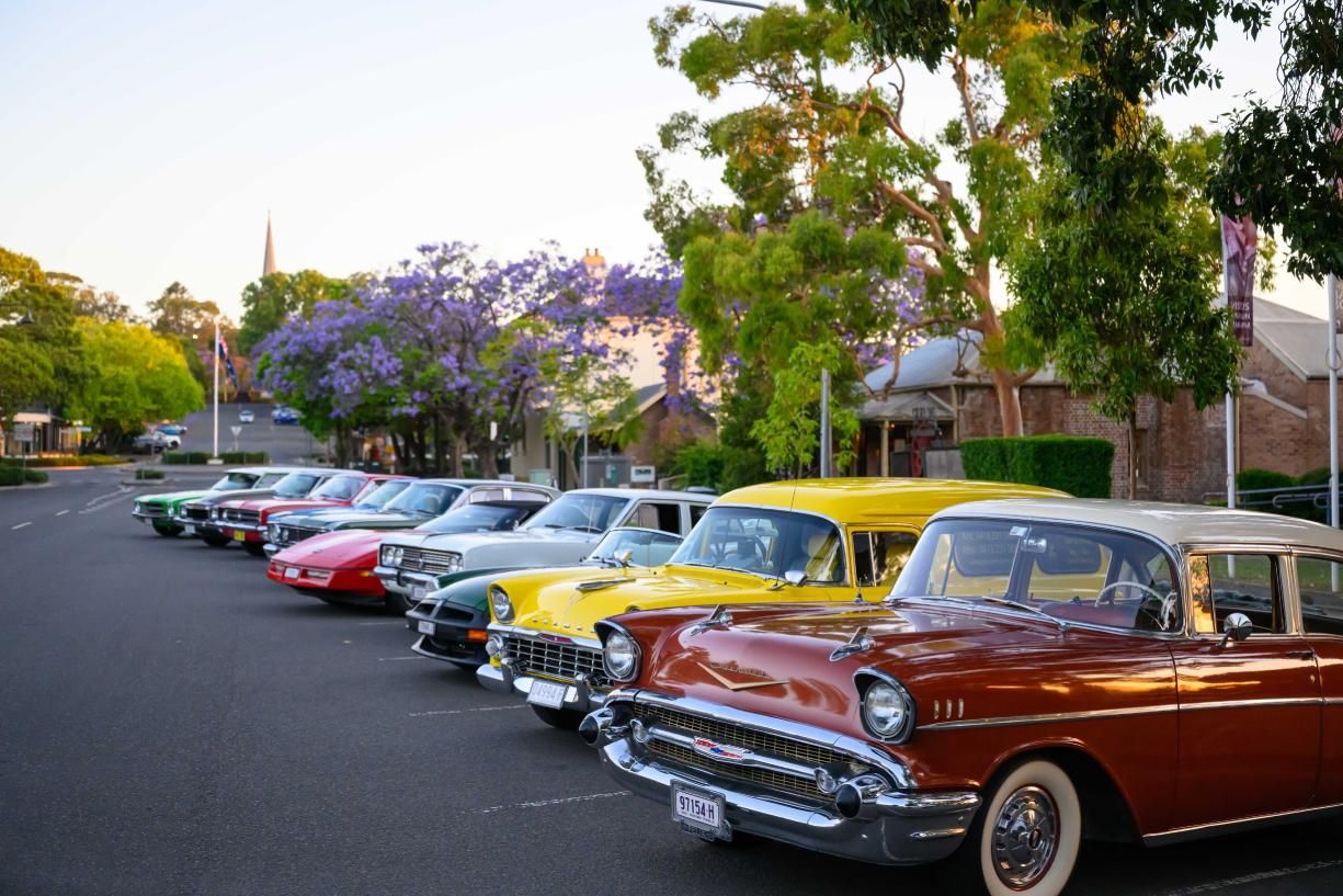 A row of vintage cars in various colors parked along a street, with trees and buildings in the background under a clear sky.