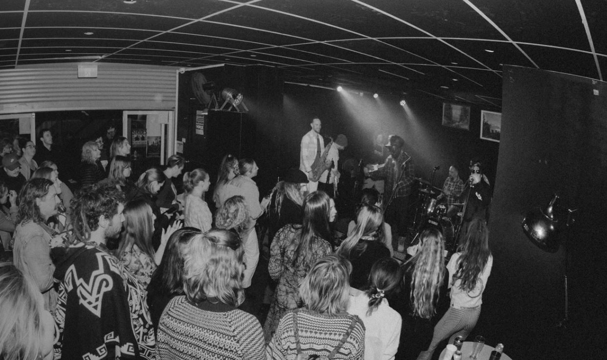 A black-and-white photo of a band performing on stage in a small venue with a crowd of people watching and enjoying the music.