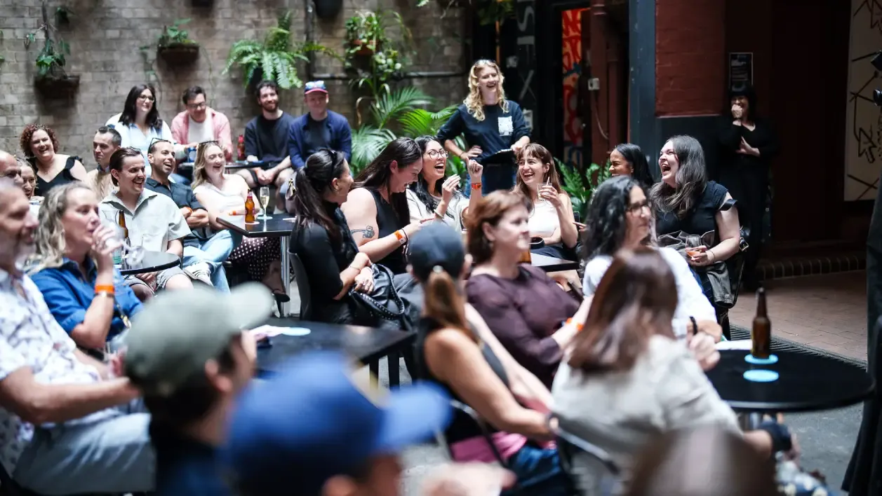A lively audience sitting at tables in a cozy venue, laughing and enjoying a live performance.
