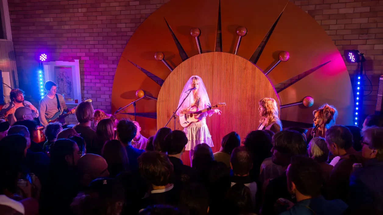 A musician in a veil performs with an acoustic guitar in front of an audience, against a backdrop featuring a large sunburst design.