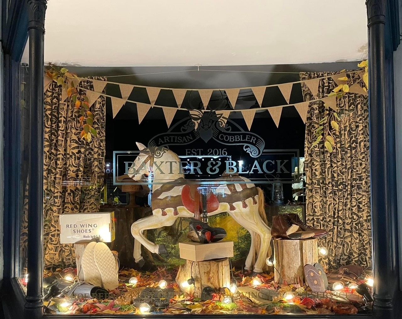 Shop window display with a fall theme, featuring a rocking horse, shoes, leaves, lights, and a banner. "Baxter & Black" sign is visible.