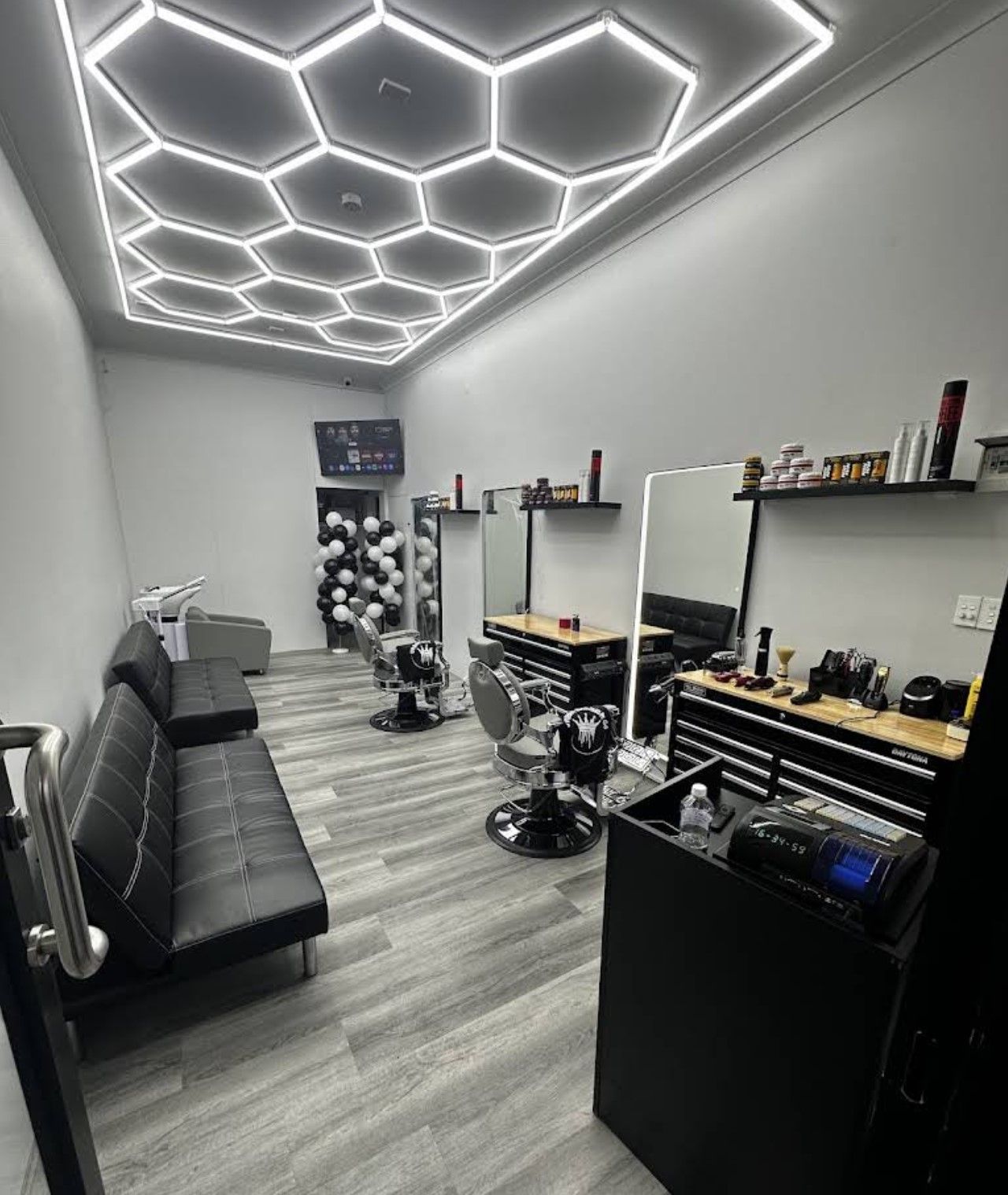 Modern barbershop with hexagonal ceiling lights, mirrors, leather chairs, tools, products, and a black leather sofa on wood flooring.