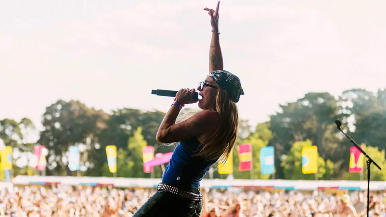 A performer with long hair and sunglasses sings passionately into a microphone on an outdoor stage, with a cheering crowd and colorful flags in the background.