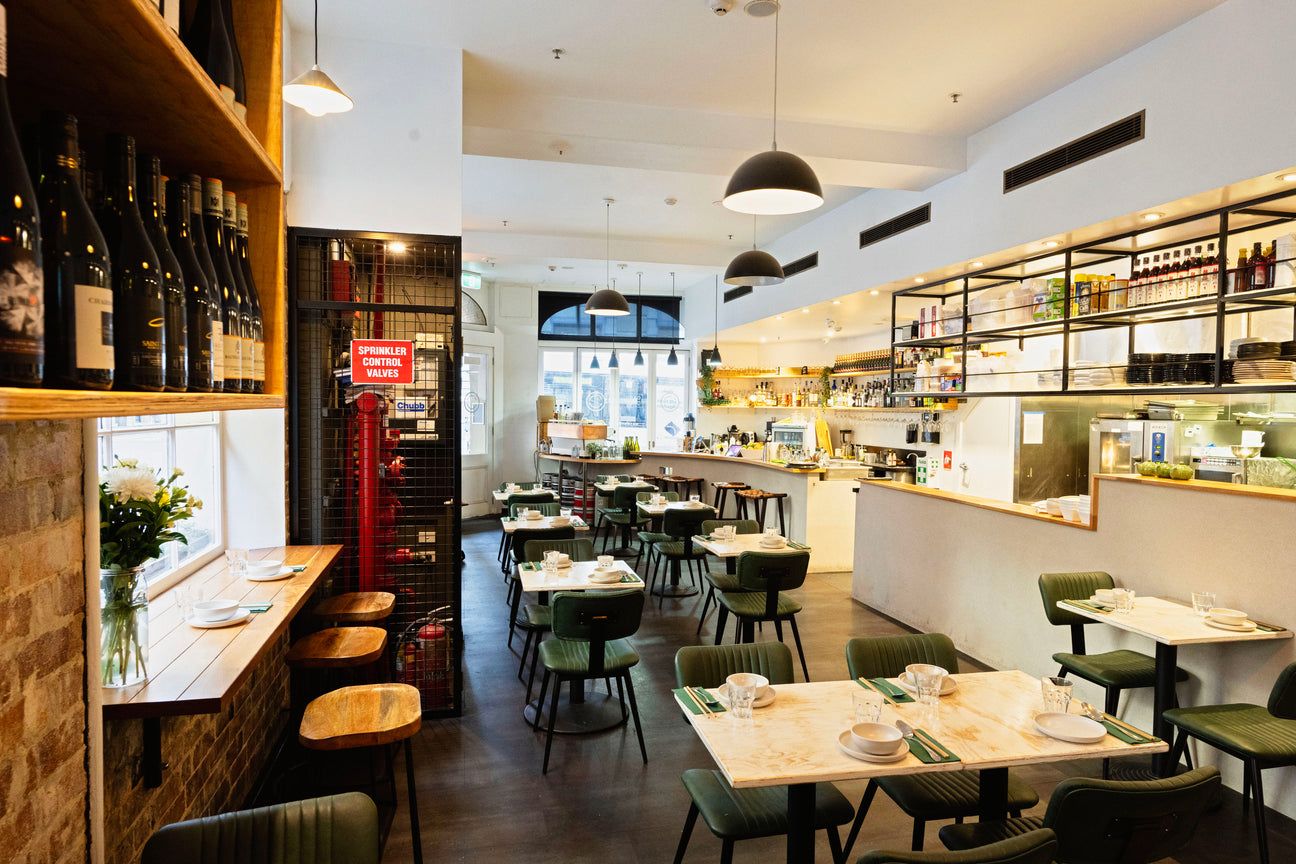Modern cafe interior with neatly arranged tables, green chairs, a bar area, wine bottles on shelves, and a "Sprinkler Control Valves" sign.
