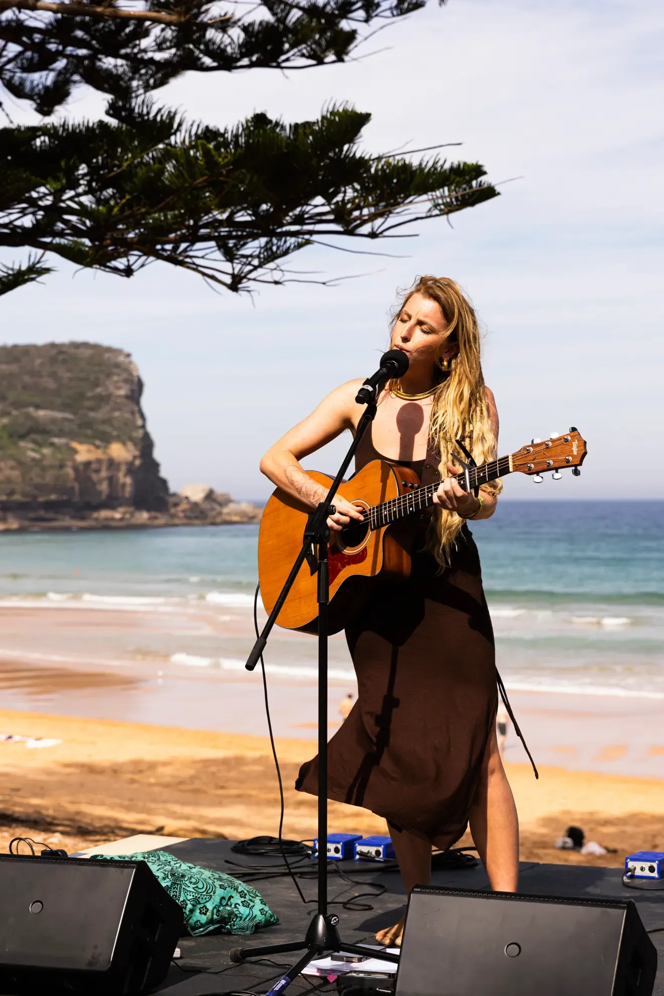 Woman playing guitar and singing on a beach stage, with ocean waves and a rocky cliff in the background, under a clear sky.