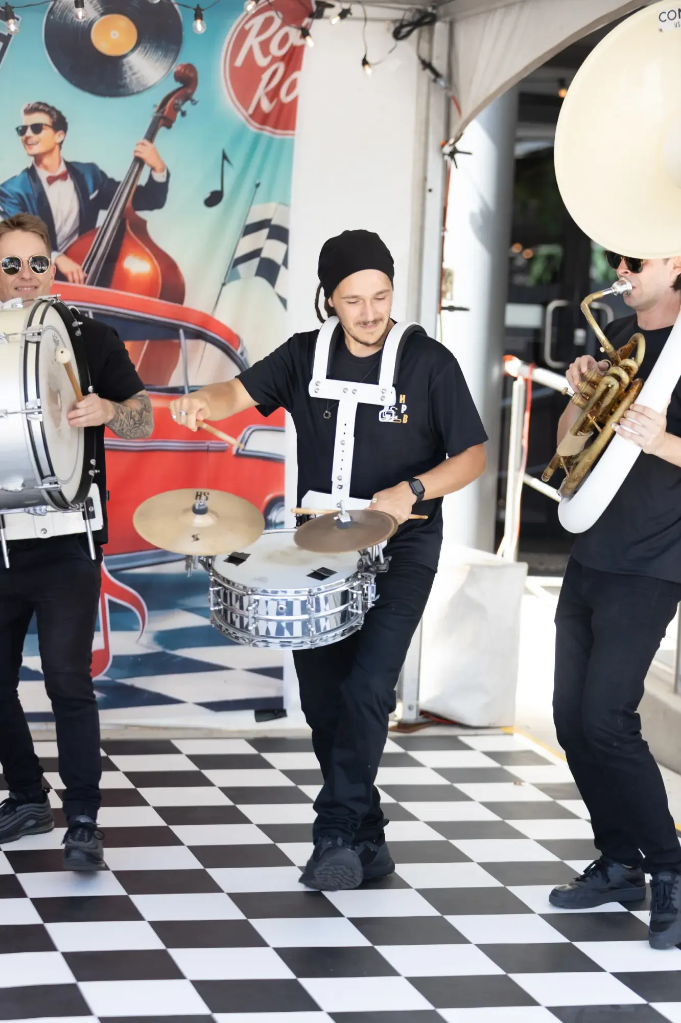 A lively trio of street musicians plays drums and tuba on a checkered floor, with a vintage car mural behind them.