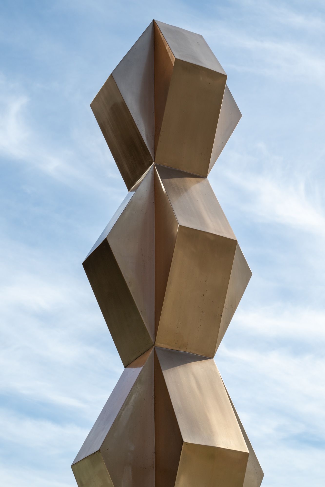 Close-up of a bronze geometric sculpture made of stacked faceted shapes, set against a blue sky with light clouds.
