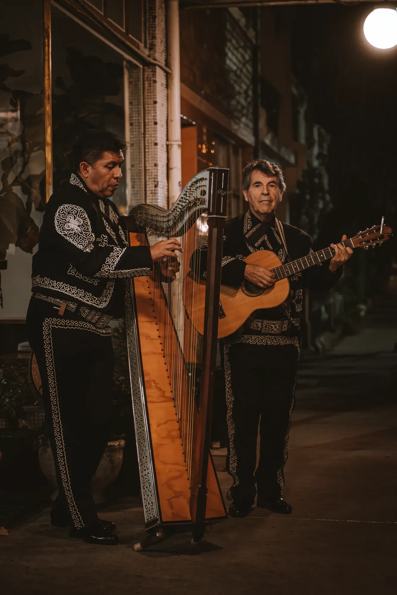 Two men in ornate black charro suits play a harp and guitar under a streetlight at night.