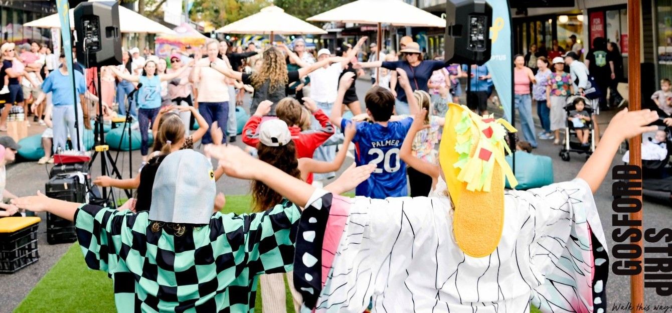 People dancing energetically at an outdoor event with performers in colourful costumes, surrounded by an audience under umbrellas.