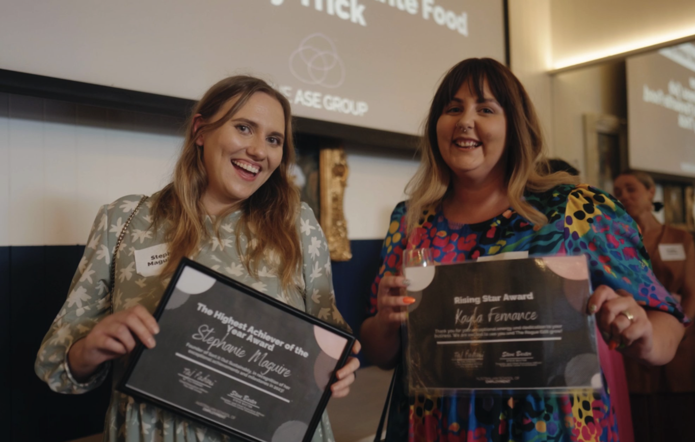 Two smiling women holding award certificates, standing in a warmly lit room with presentation slides in the background.